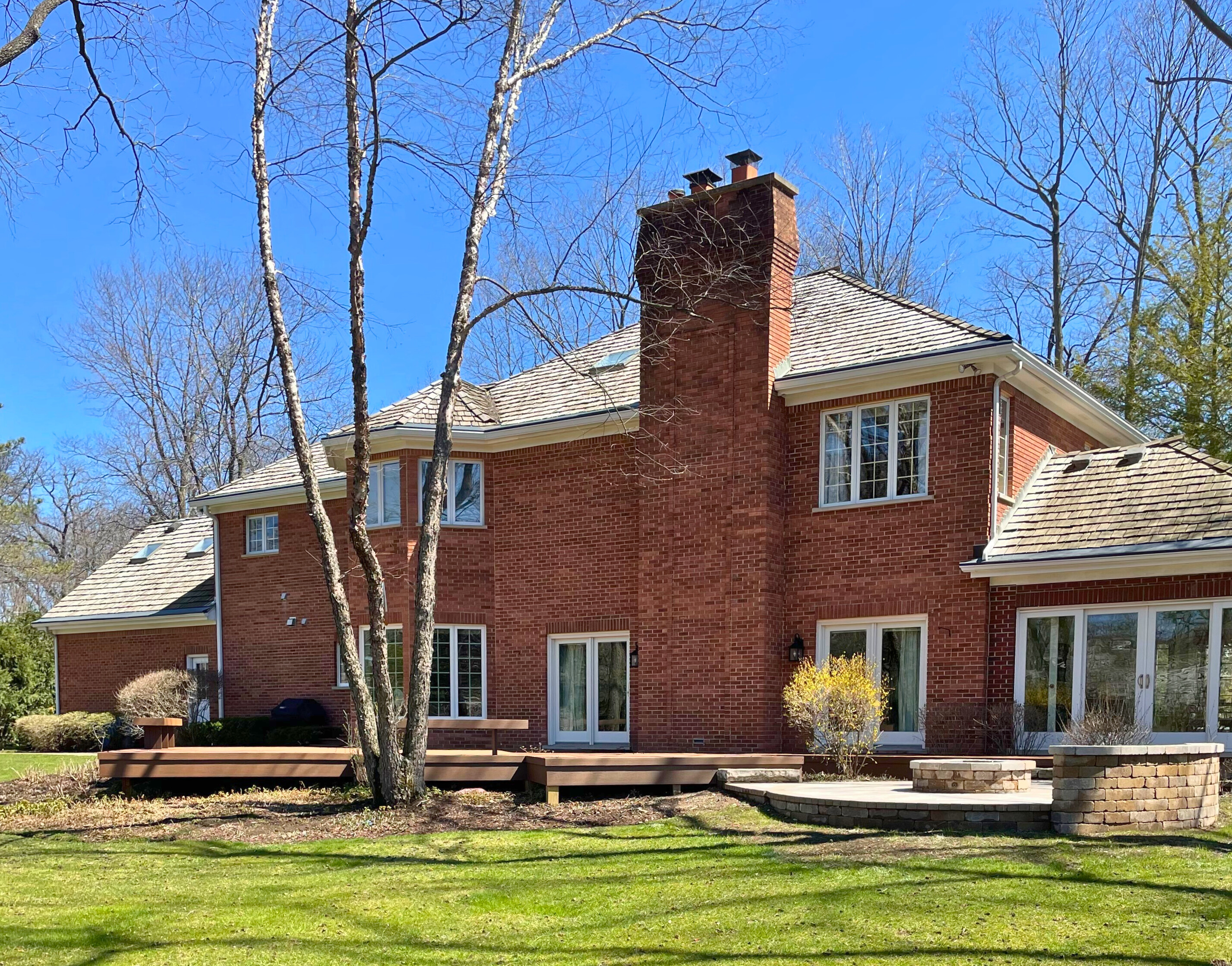 A spacious, low-profile dark composite deck extending from a large red brick home with multiple windows and doors, leading to a circular stone fire pit area on a paver patio, set against a backdrop of bare trees and a sunny sky in Lake Forest, IL.