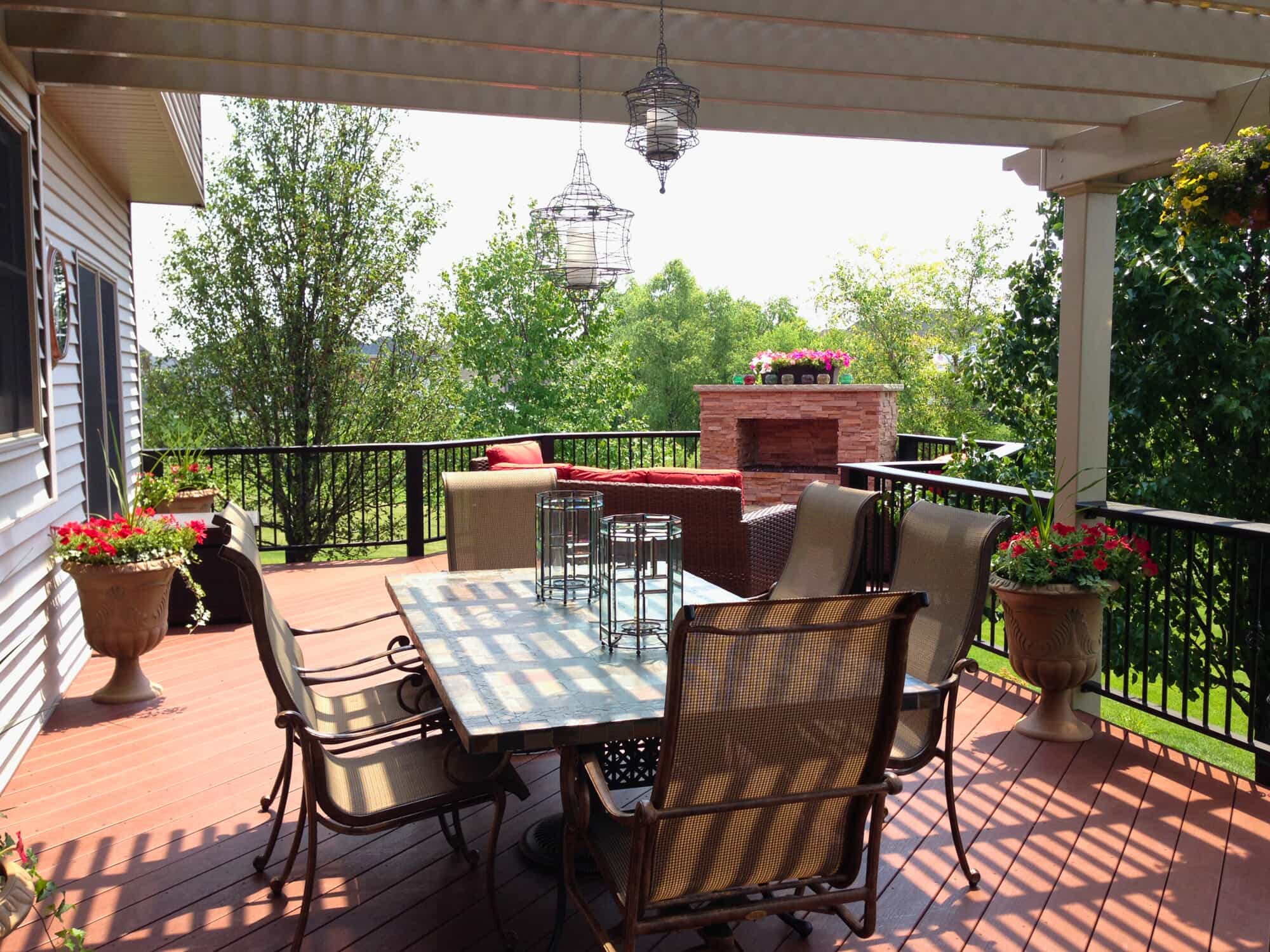 A spacious wooden deck with a dark railing and a pergola overhead, furnished with outdoor seating and a dining table. Potted flowers add splashes of color.