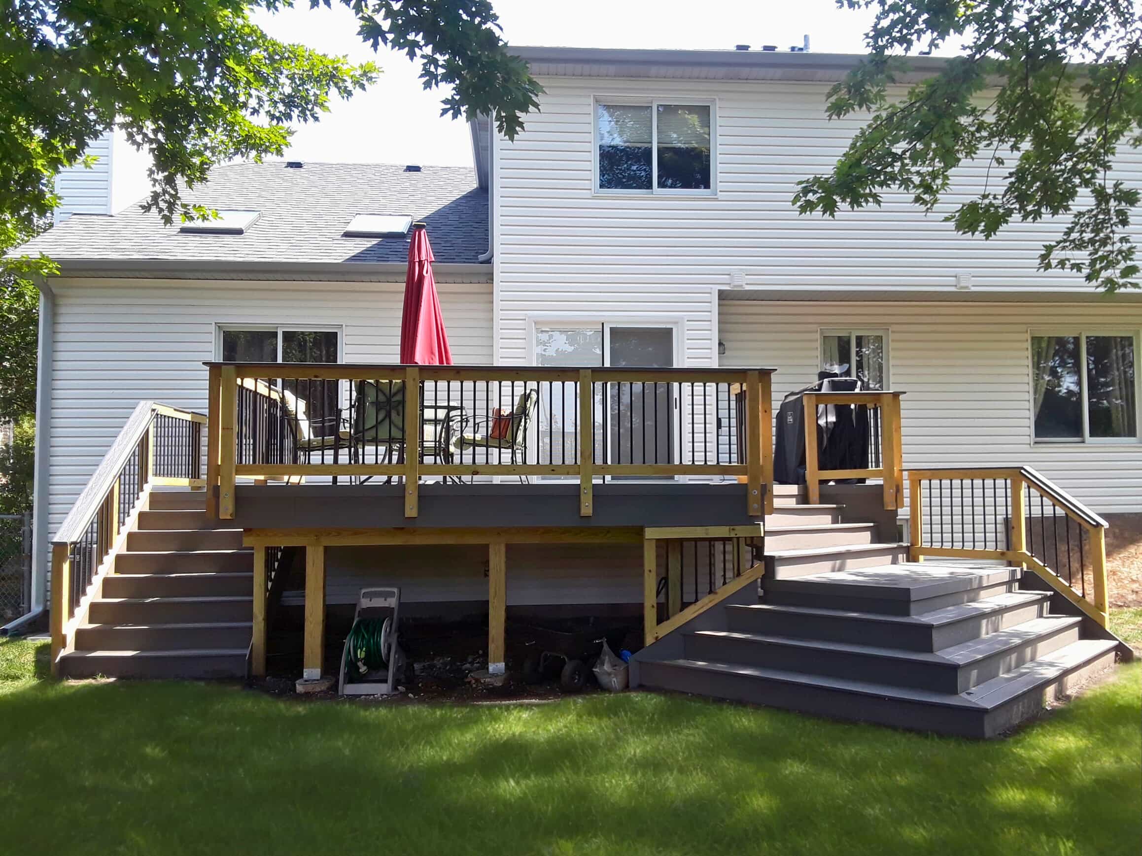 A multi-level composite deck with two sets of stairs leading down to a green lawn. The deck has dark gray flooring and natural wood railings with black balusters. An umbrella and outdoor furniture are visible on the upper level.