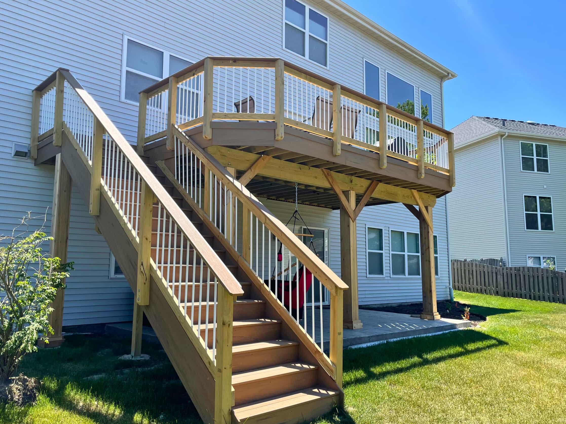 A raised wooden deck attached to a light-colored house, with a long set of stairs leading down to a green lawn. The deck has a natural wood finish with white balusters