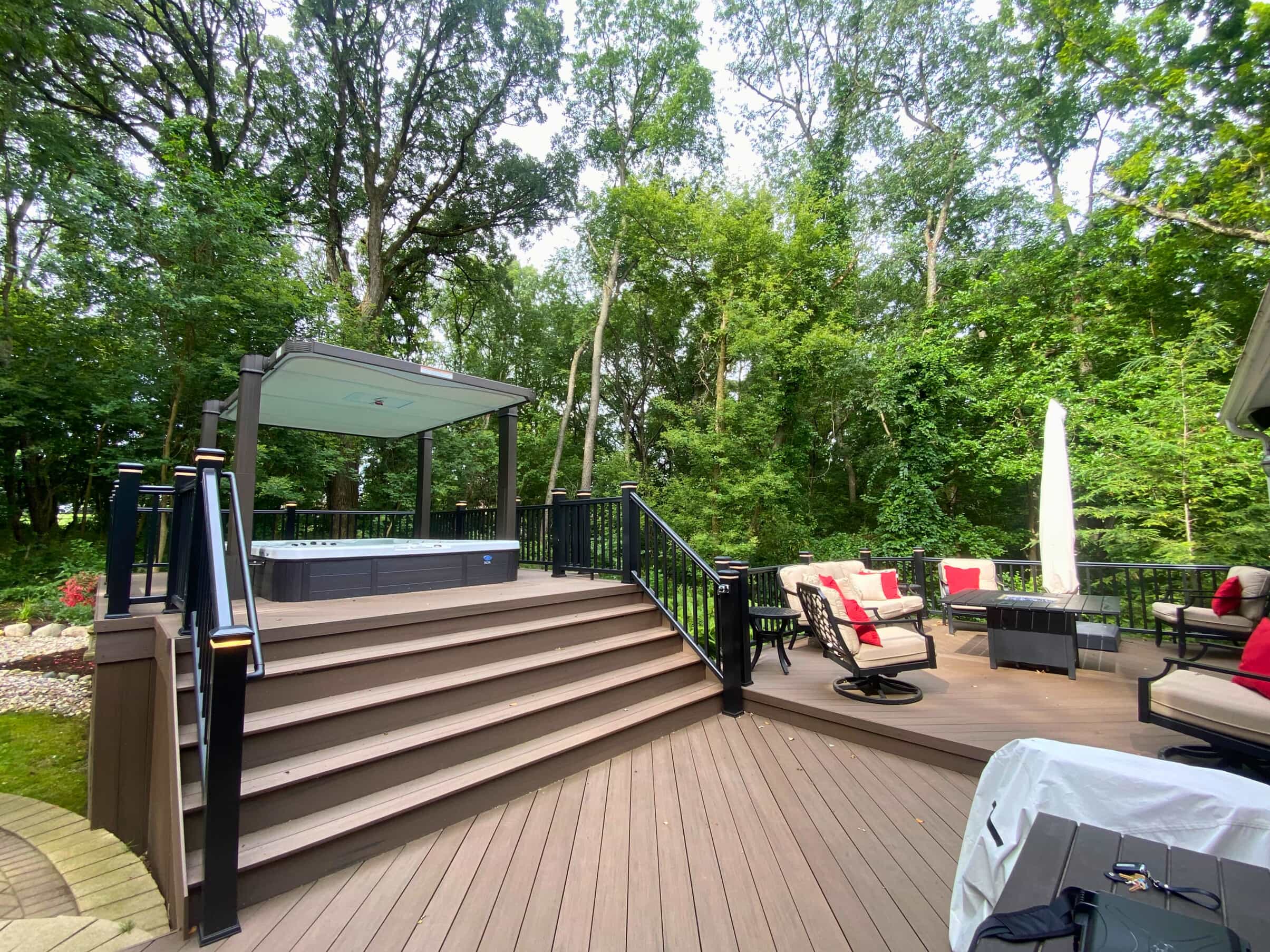 Custom multi-level composite deck in St. Charles, IL, with black railings, integrated hot tub, and a pergola over the hot tub, surrounded by lush trees.