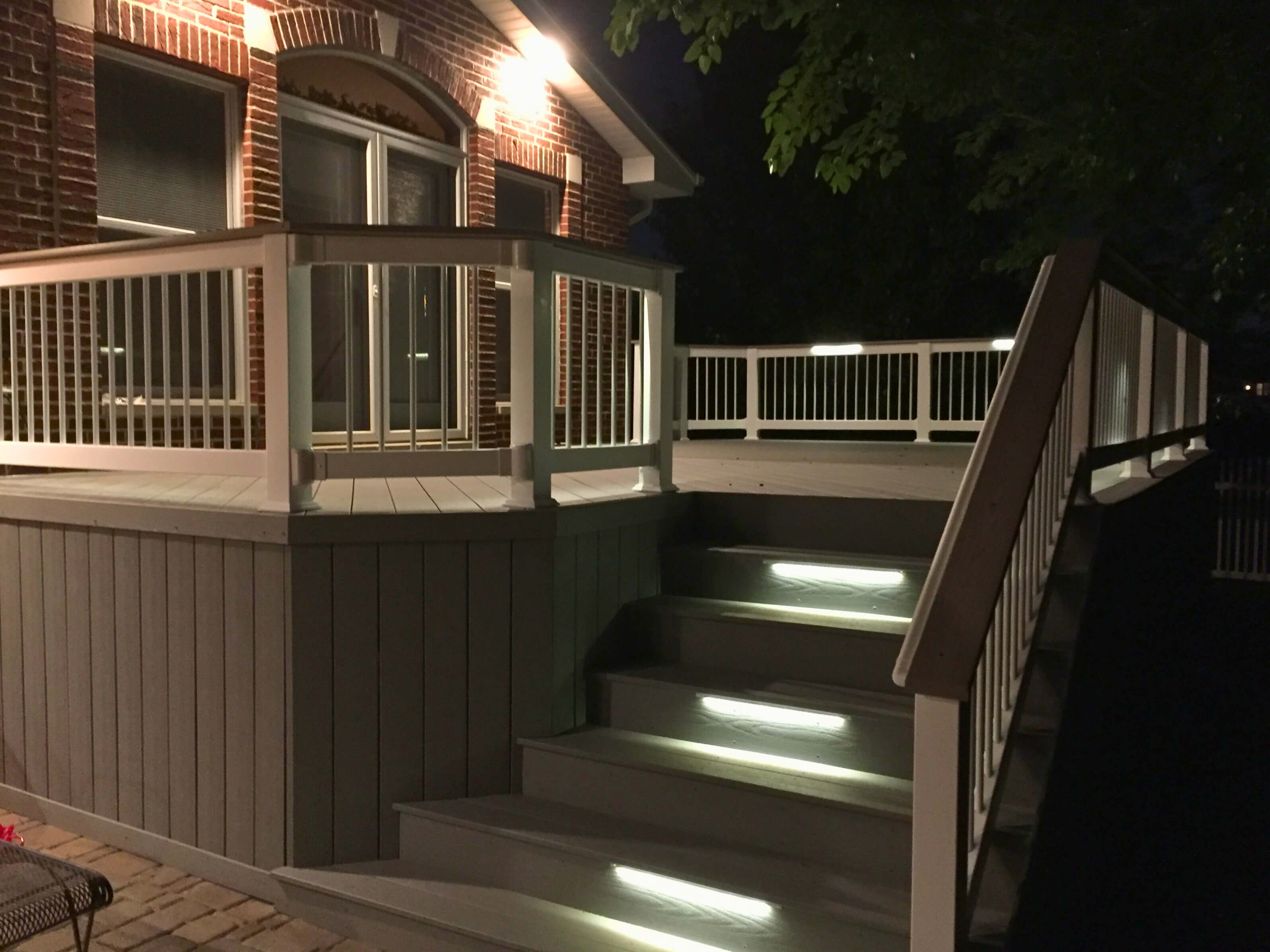 Illuminated composite deck with white railings and stairs leading down to a patio at night, showcasing a brick house in Tinley Park, IL.
