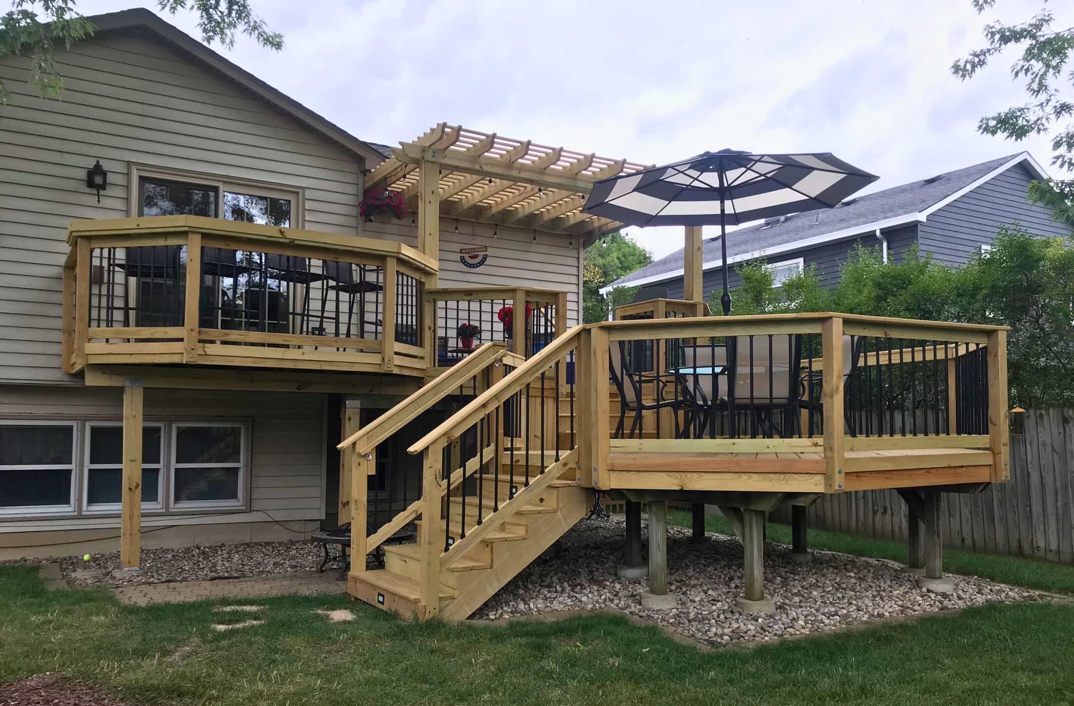 A multi-level wooden deck with two distinct sections, one featuring a small pergola. The deck has natural wood railings with black balusters and steps leading down to a landscaped area with rocks.