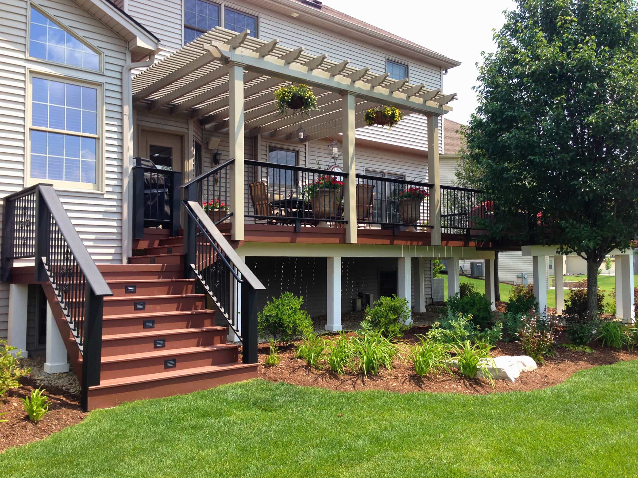 A large elevated deck with a wooden pergola covering part of it. The deck has dark reddish-brown flooring and black railings, with stairs leading down to a well-maintained lawn and garden.