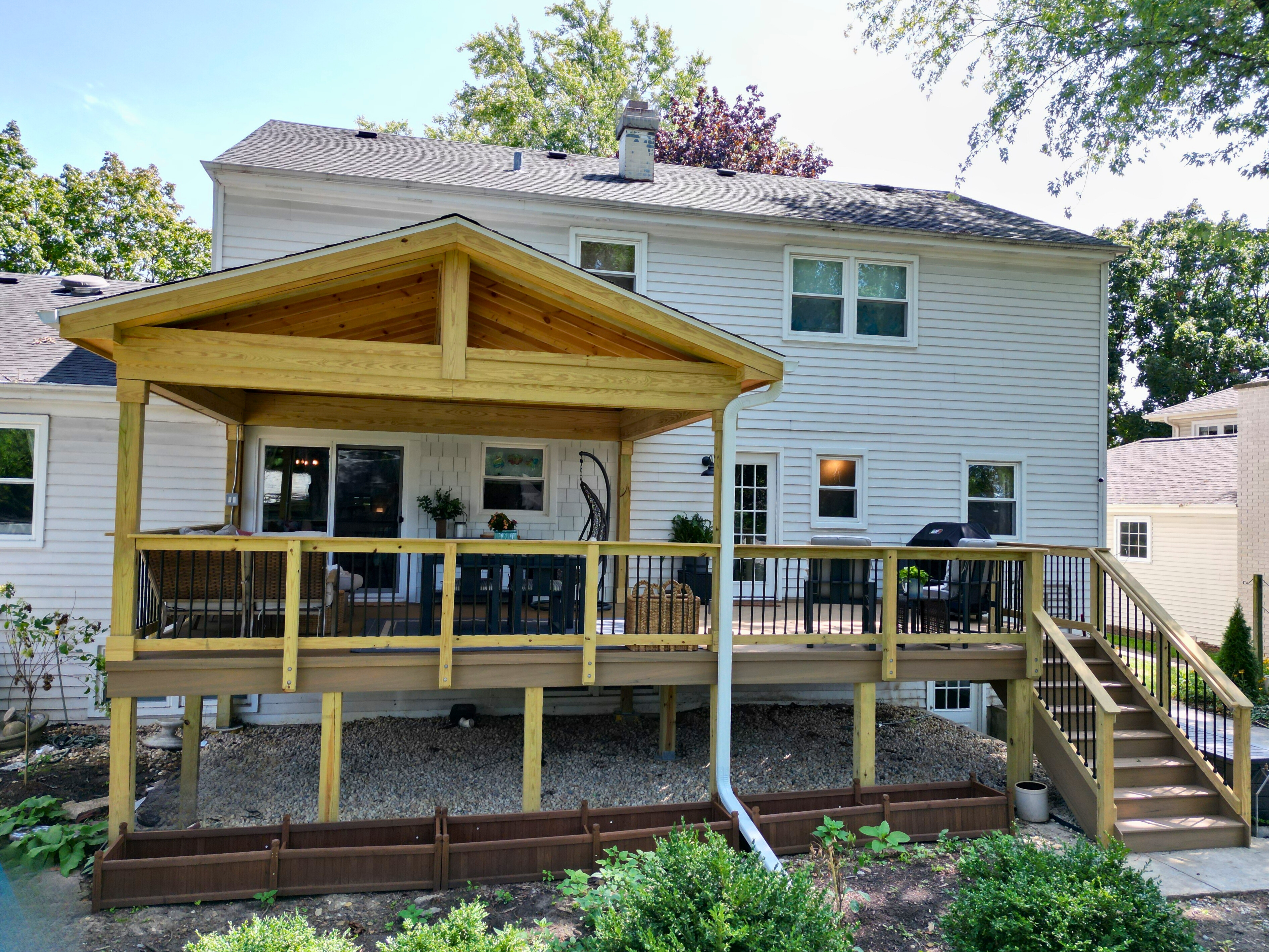 Custom covered deck with a gable roof and natural wood ceiling, extending from a white house in Downers Grove, IL. Built by Archadeck of Chicagoland, this deck features brown decking, black railings, and provides shaded outdoor living.