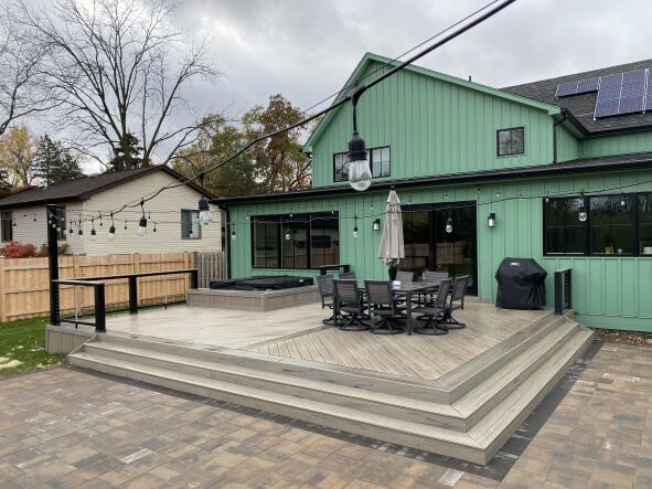 Modern multi-level composite deck in shades of gray, extending to a paver patio, with string lights overhead and a hot tub, built for a green house in Downers Grove, IL by Archadeck of Chicagoland.