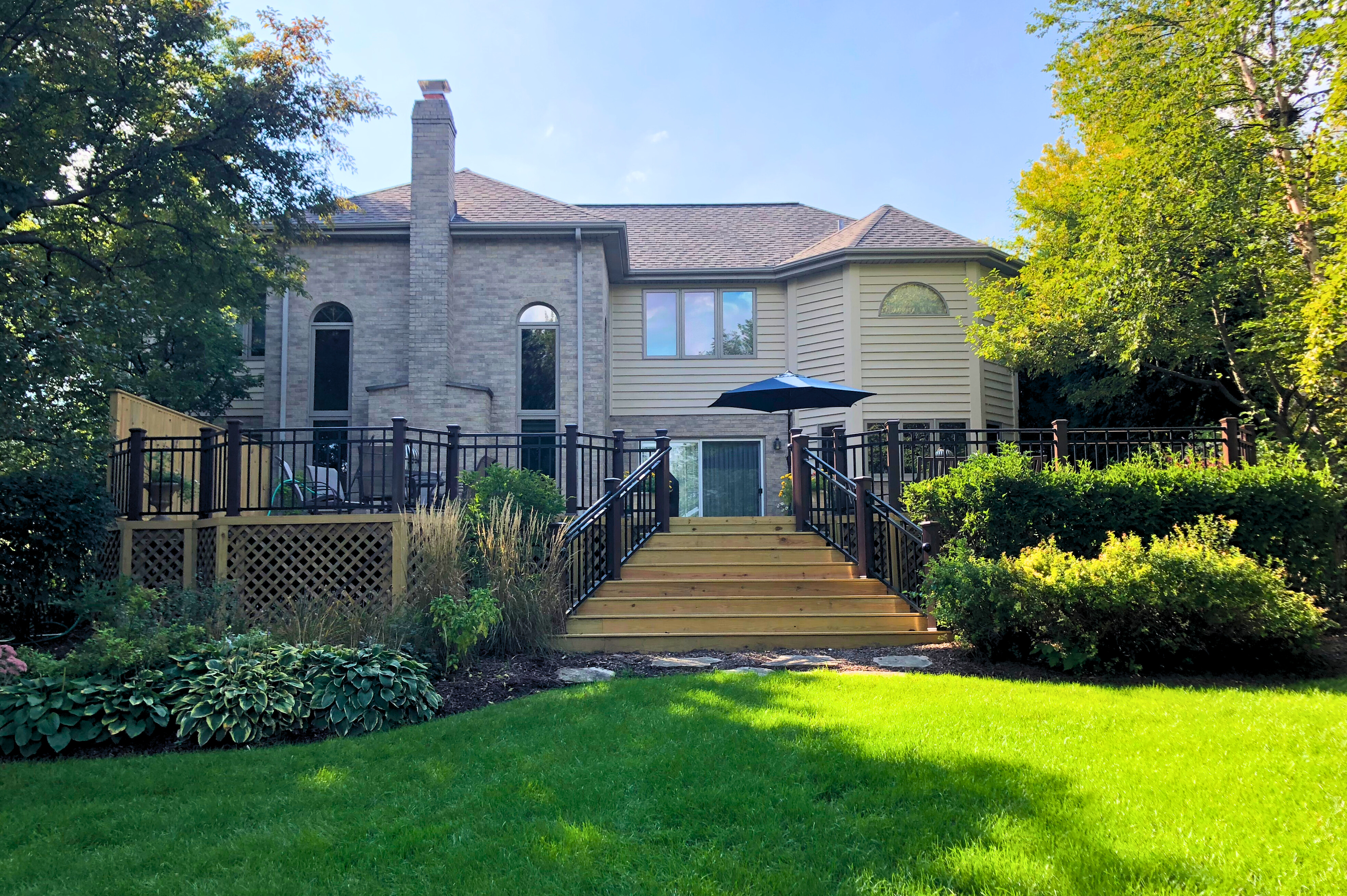 Large custom wood deck with black metal railings and wide steps leading to a green lawn, built for a brick and siding house in Downers Grove, IL. A beautiful example of a deck by Archadeck of Chicagoland.