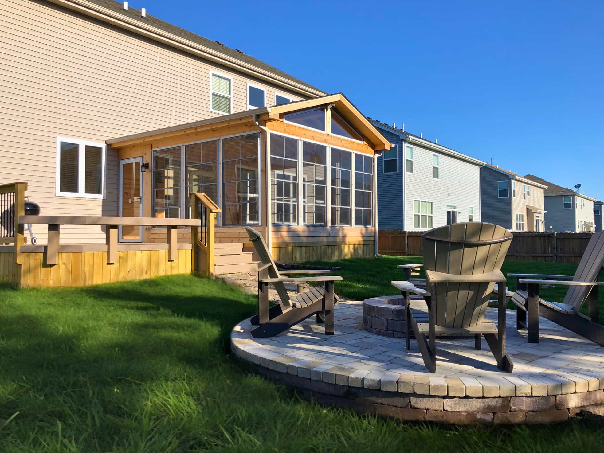 A house with a light-colored exterior and a large sunroom addition. To the left is a wooden deck, and in the foreground, a paver patio with a circular fire pit and four chairs is visible on a green lawn.
