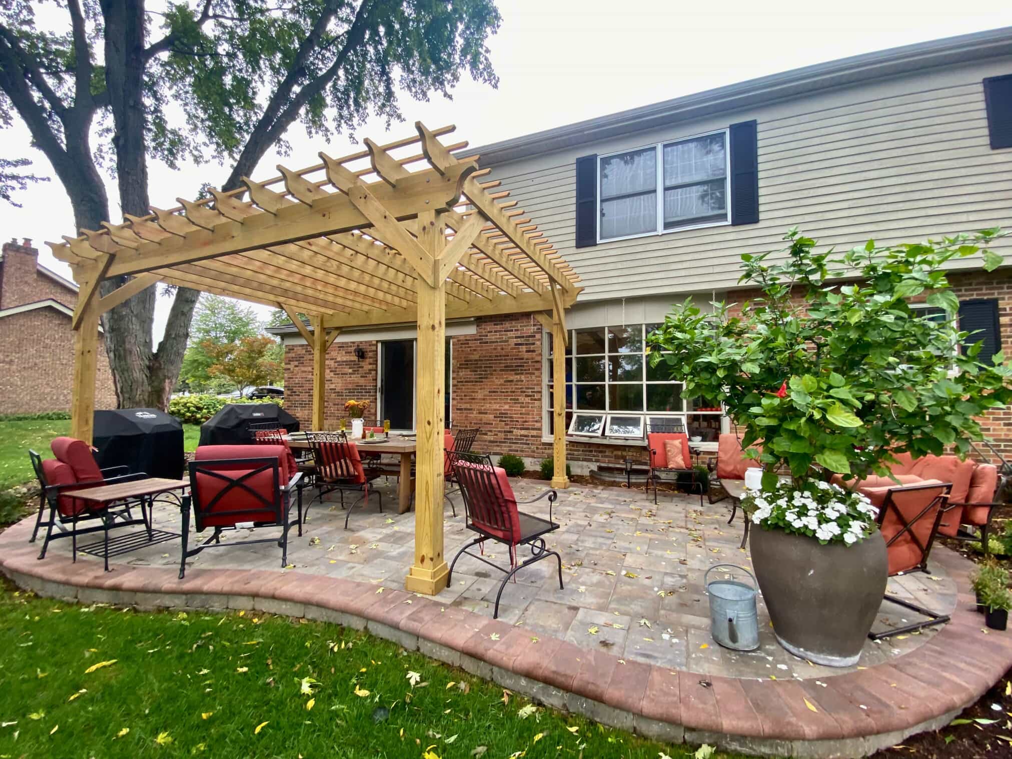 Outdoor paver patio in St. Charles, IL, with a wooden pergola covering a dining set, next to a brick house and lush greenery.