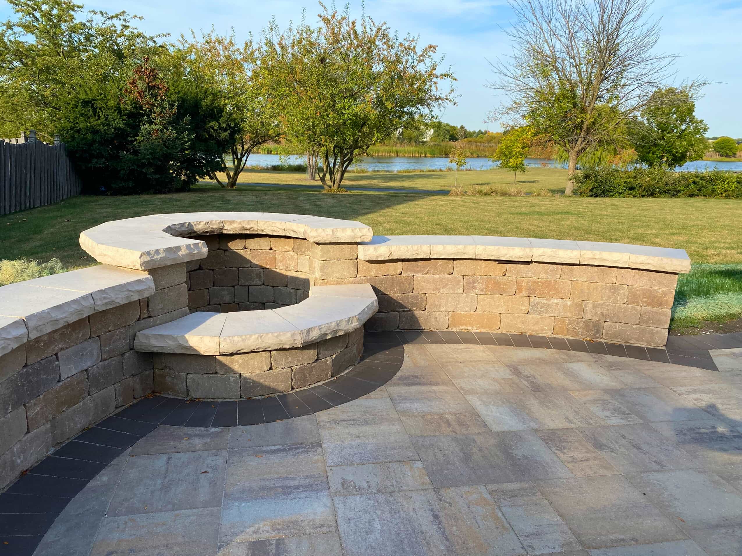 A beautifully constructed paver patio with a built-in curved seating wall and a fire pit. The patio features varying shades of stone and is surrounded by a green lawn with trees and a lake in the background.