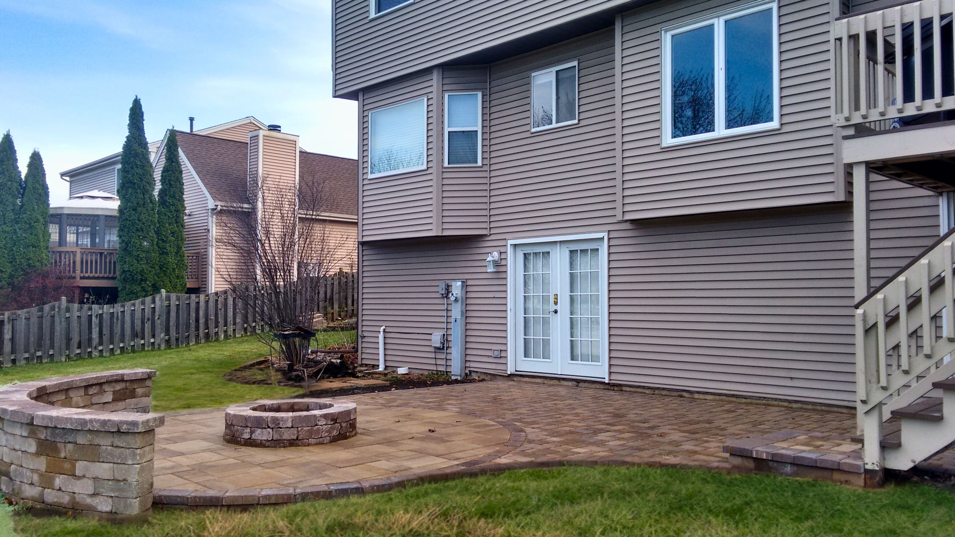Circular paver patio in St. Charles, IL, featuring a built-in stone fire pit and a matching curved stone seating wall, next to a house with a deck staircase.
