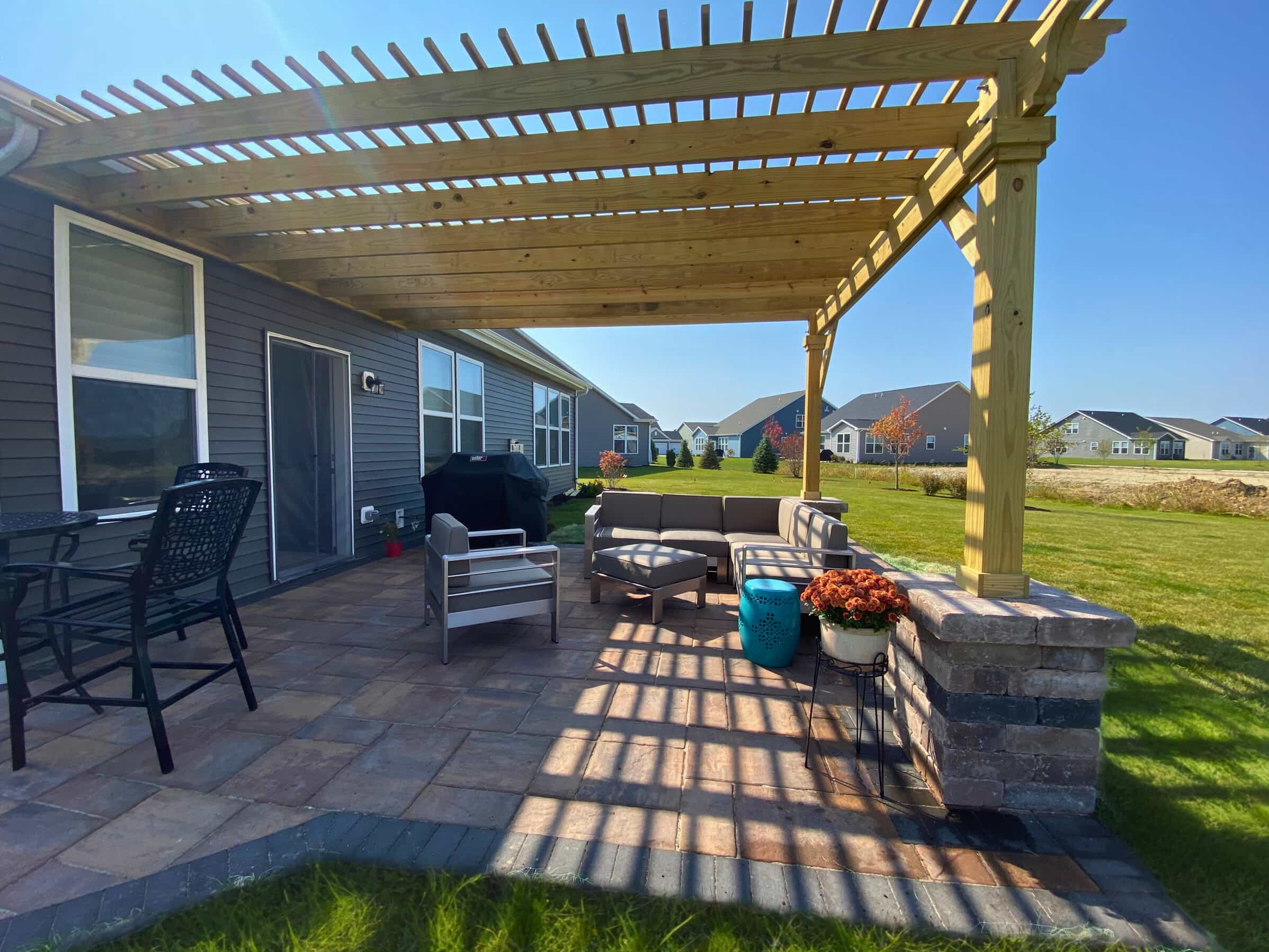 A large wooden pergola extending over a paver patio with outdoor furniture. The pergola has a natural wood finish and is supported by stone pillars.