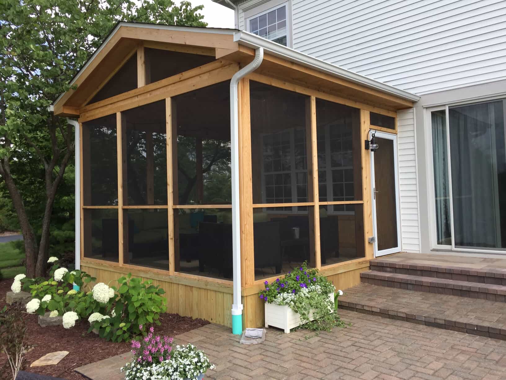 Exterior view of a newly constructed screened porch addition on a light-colored house, featuring natural wood framing, dark screening, and a brick patio leading to it.
