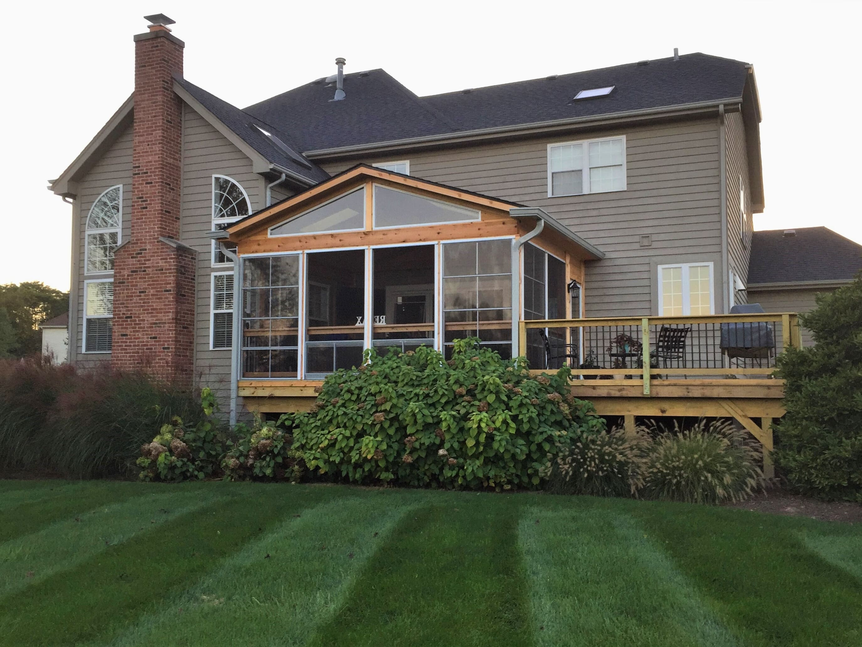 Exterior view of a large screened porch addition in St. Charles, IL, with a gable roof and an attached wooden deck, next to a two-story house.