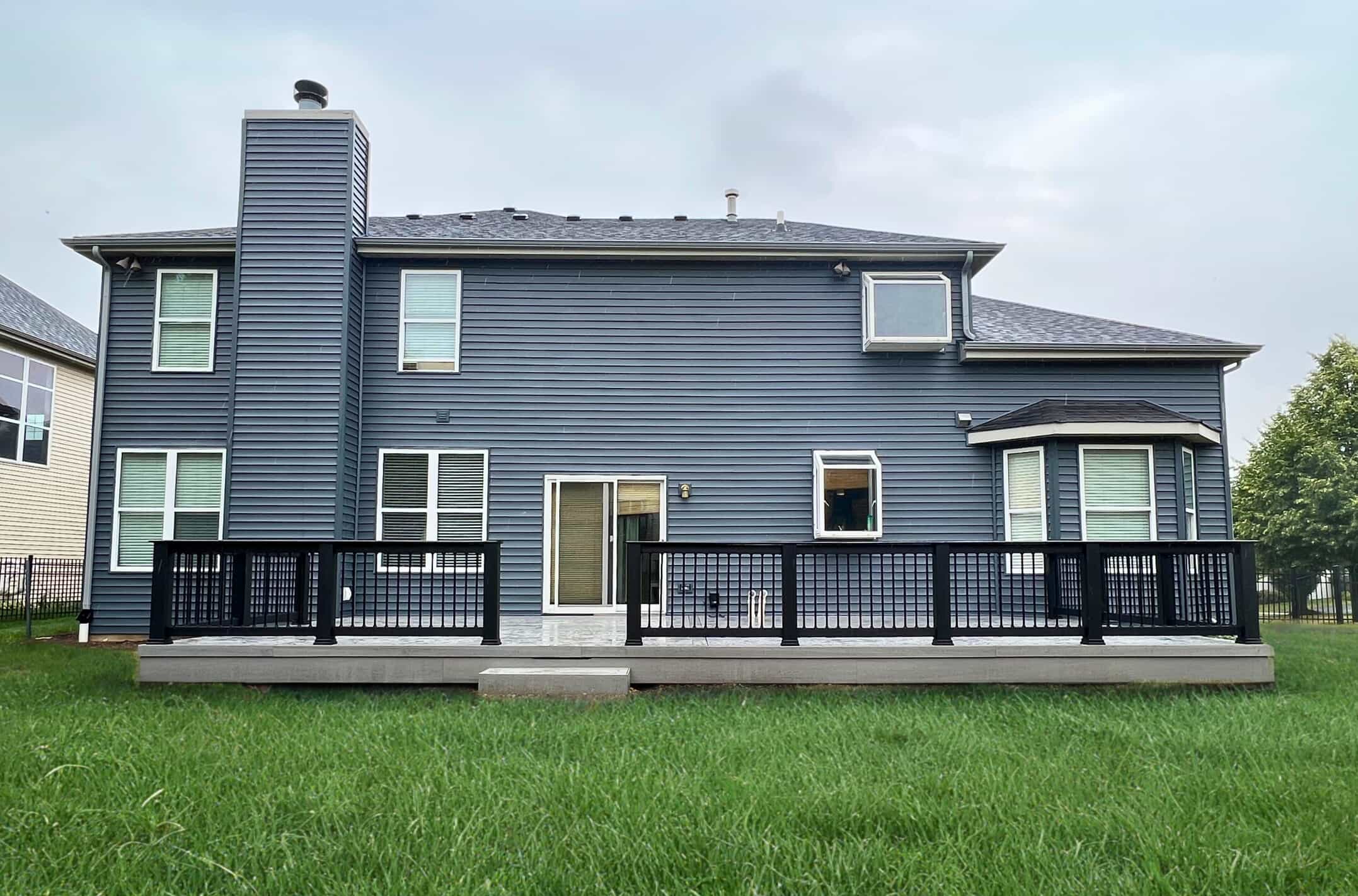 The back of a gray house with a wide, low-profile TimberTech composite deck. The deck has dark gray flooring and black railings, extending across the entire back of the house.