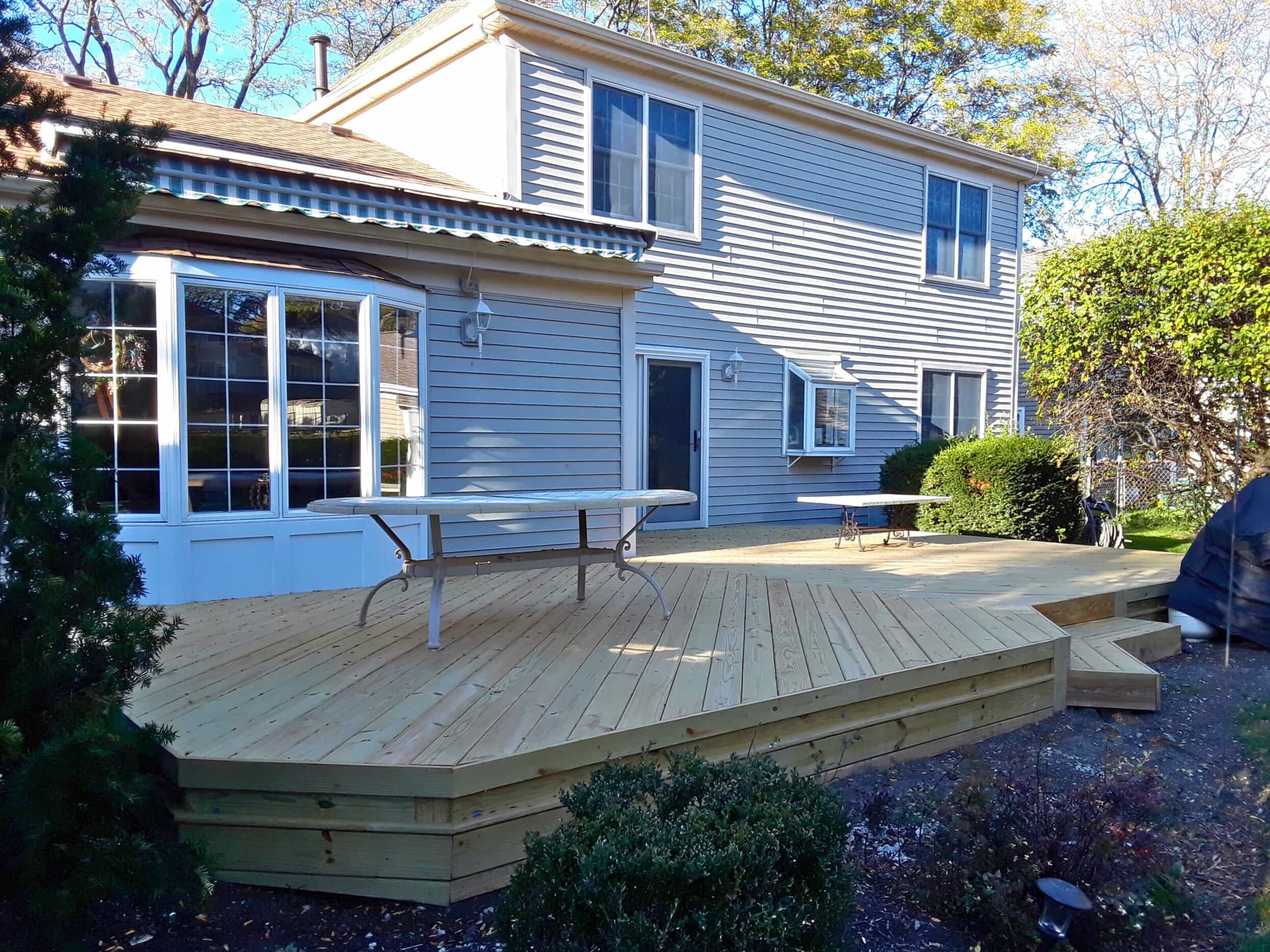 Large, newly built platform wood deck with steps, extending from a light blue siding house with a bay window in Tinley Park, IL.