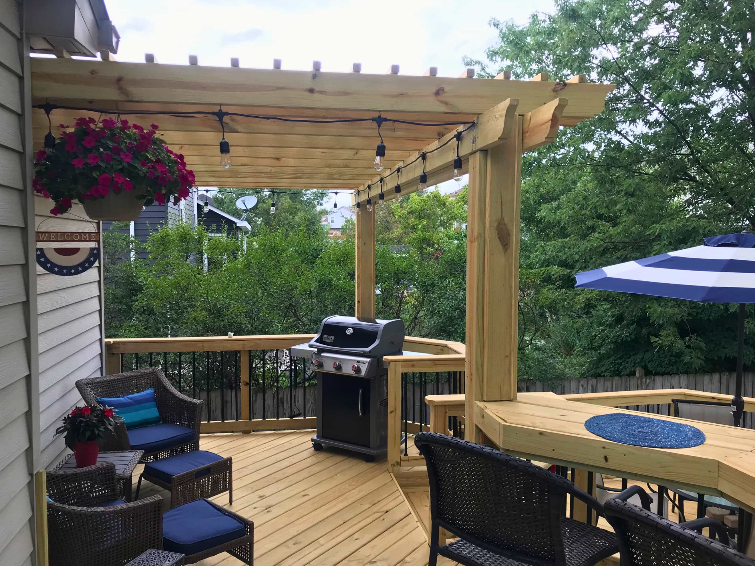 A spacious natural wood deck with a pergola covering part of it, featuring string lights, outdoor furniture, a grill, and a built-in bar counter. A hanging flower basket and a welcome sign are visible near the house.