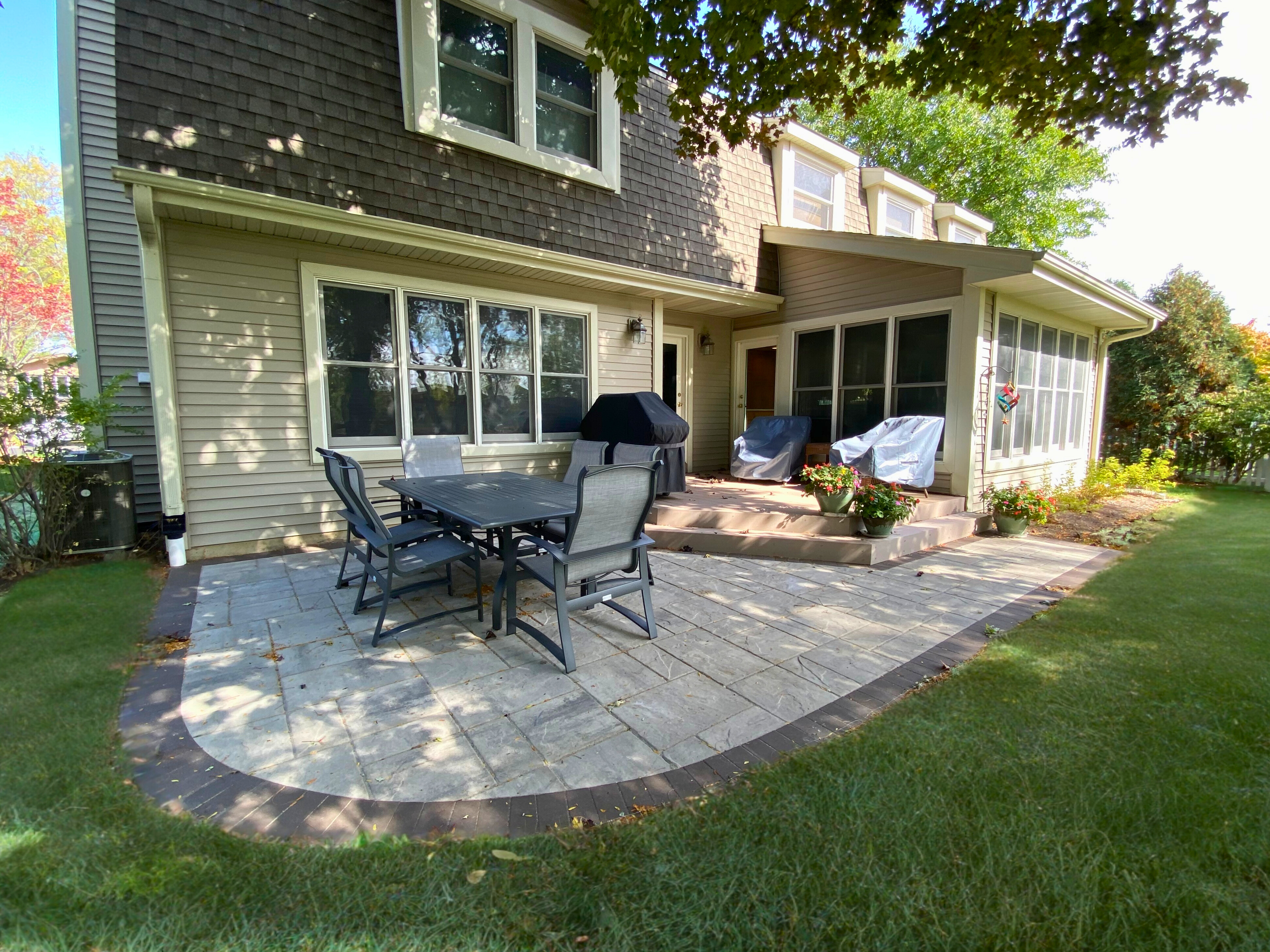 Large curved paver patio behind a house in Wheaton, IL. The patio features light gray pavers and connects to a sunroom, with outdoor furniture and a grill.