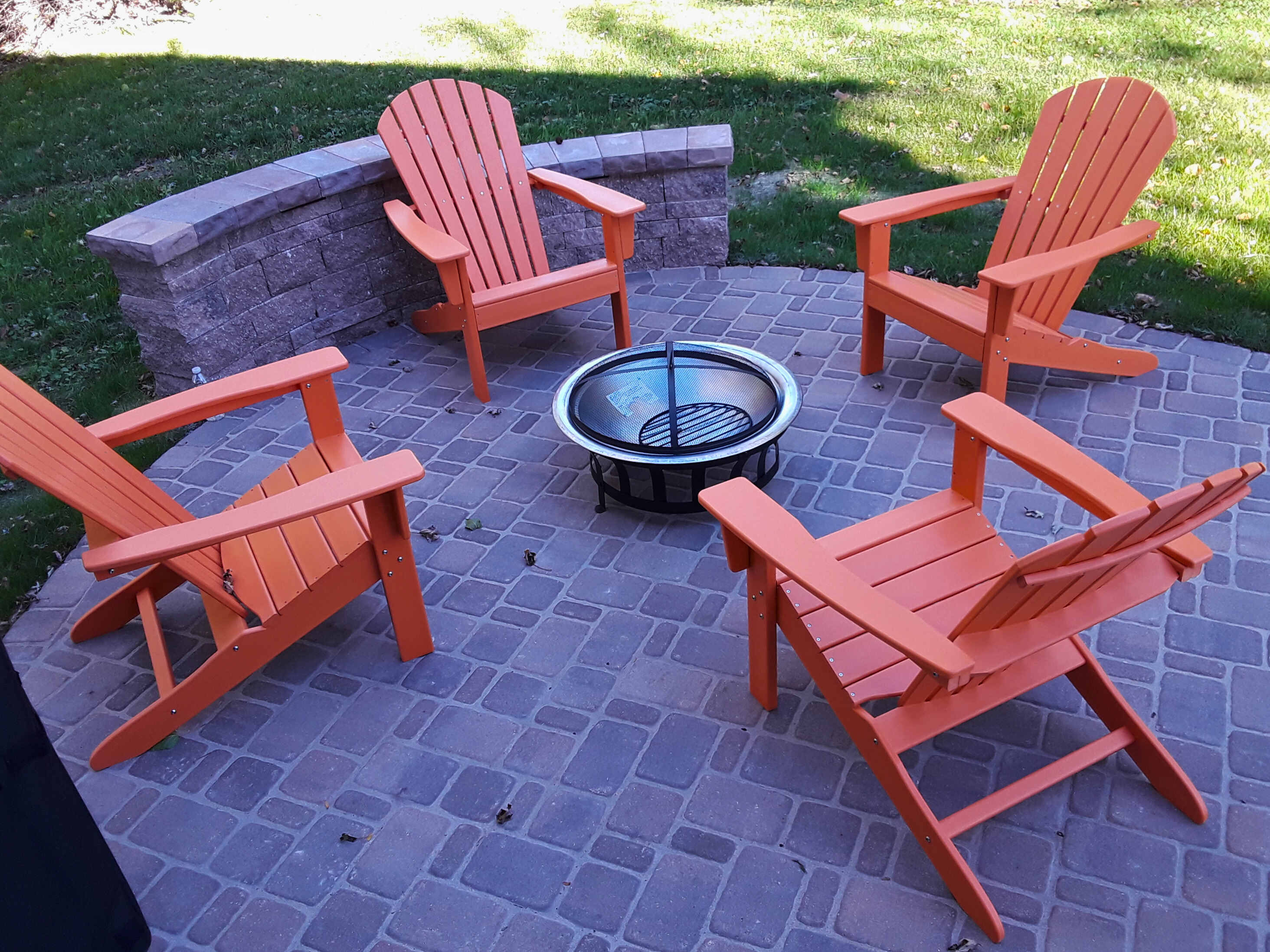 Paver patio in Wheaton, IL with a built-in stone seating wall, a metal fire pit, and four orange Adirondack chairs. Ideal for outdoor gatherings and warmth.