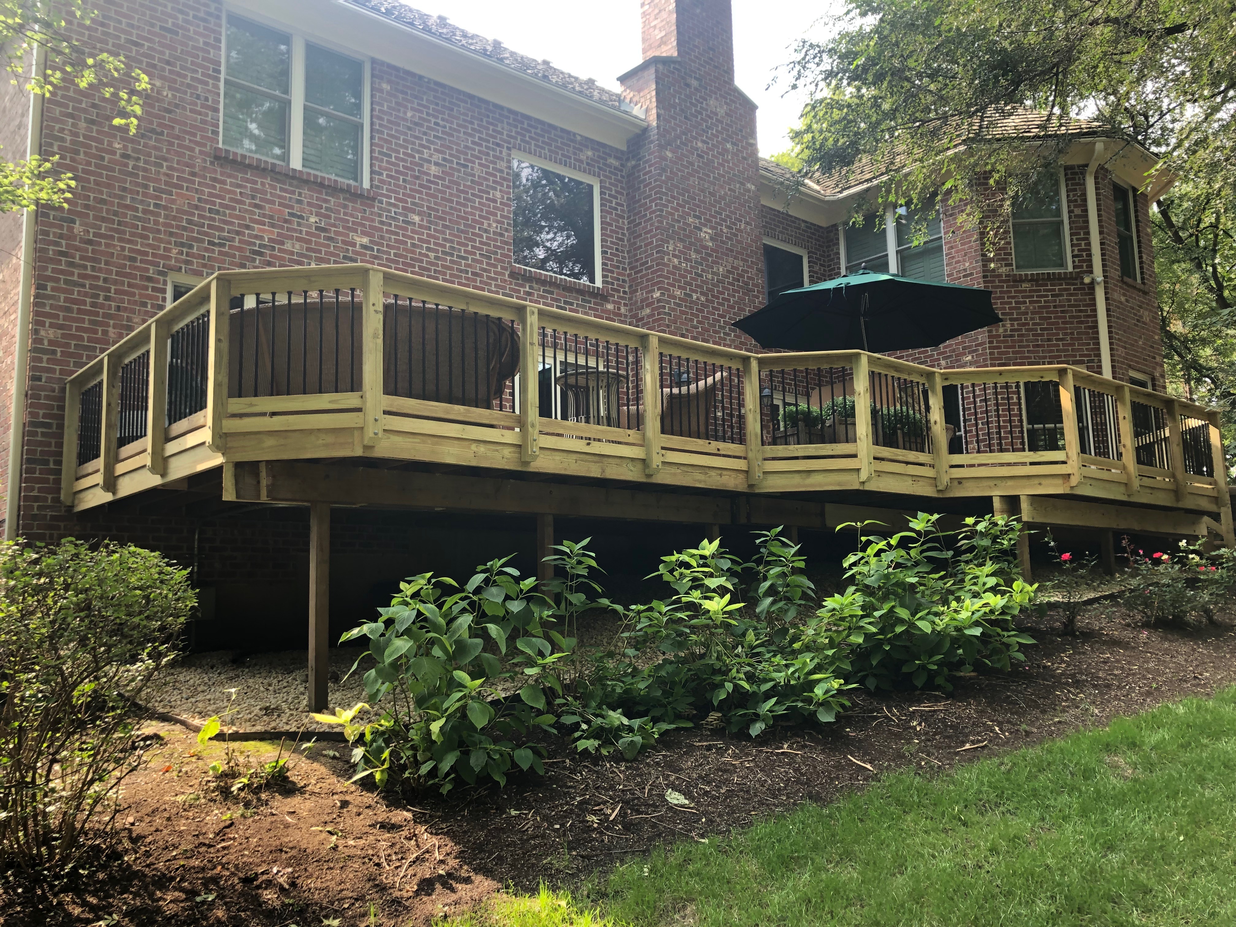 A large elevated wooden deck with black balusters and natural wood railings, featuring a built-in hot tub, attached to a brick home with multiple windows, overlooking a landscaped backyard with lush greenery, under a sunny sky in Lake Forest, IL.