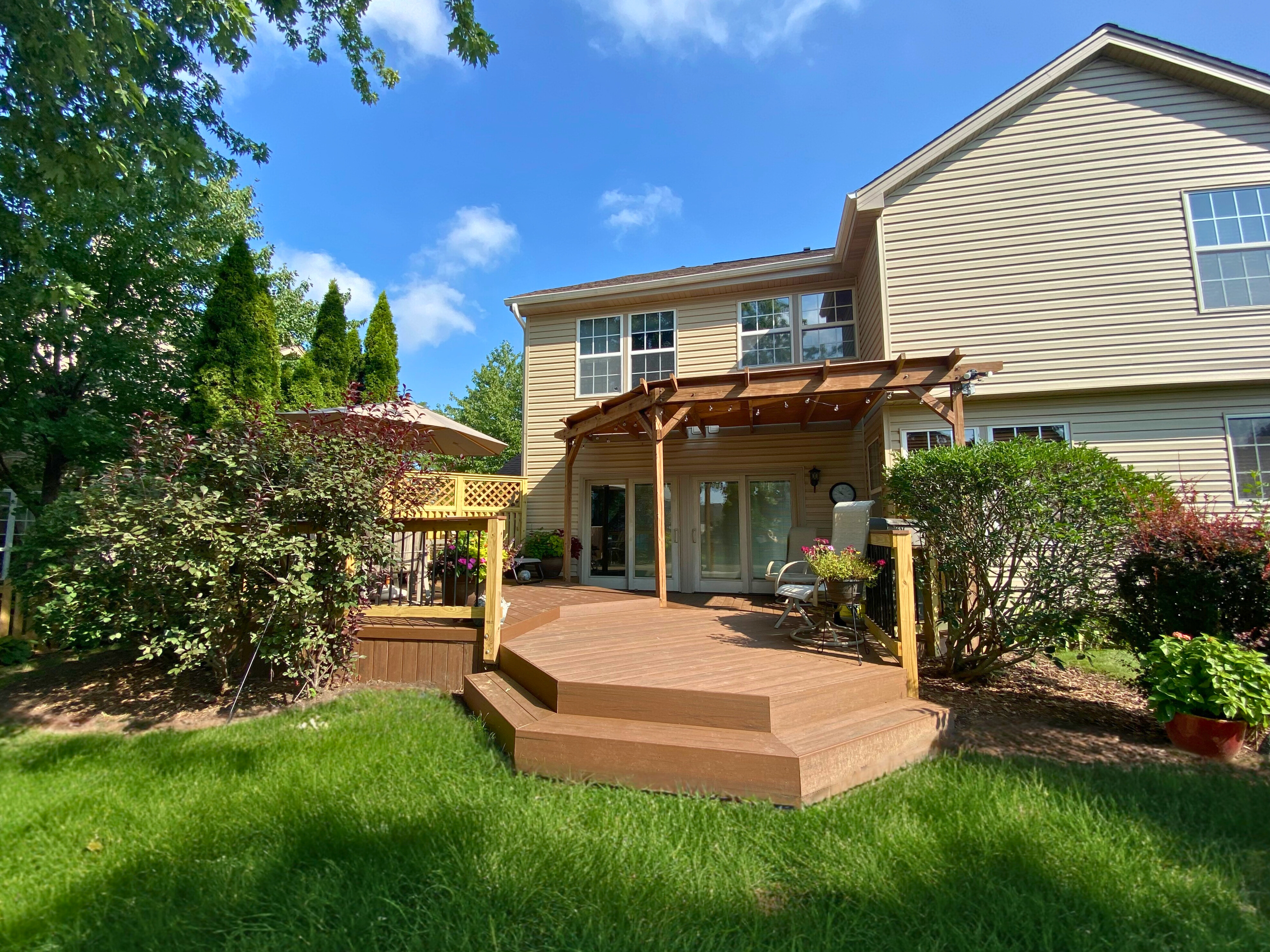 An expansive wooden deck with a black and natural wood railing, surrounding an in-ground swimming pool and furnished with various outdoor seating, all set within a lush green treed backyard.
