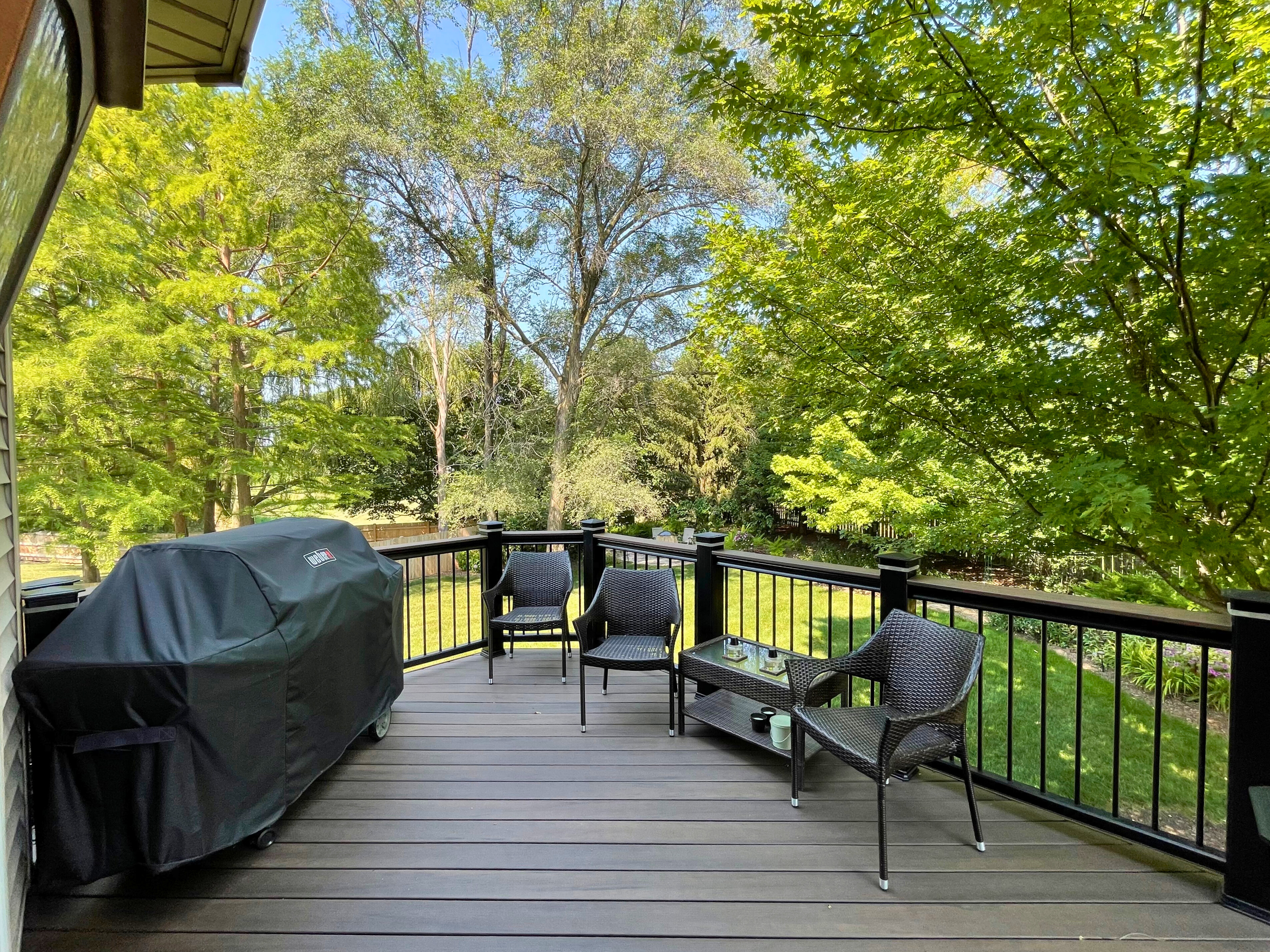 Composite deck in Mundelein, IL, featuring dark decking, black railings, outdoor furniture, and a grill, surrounded by lush trees.