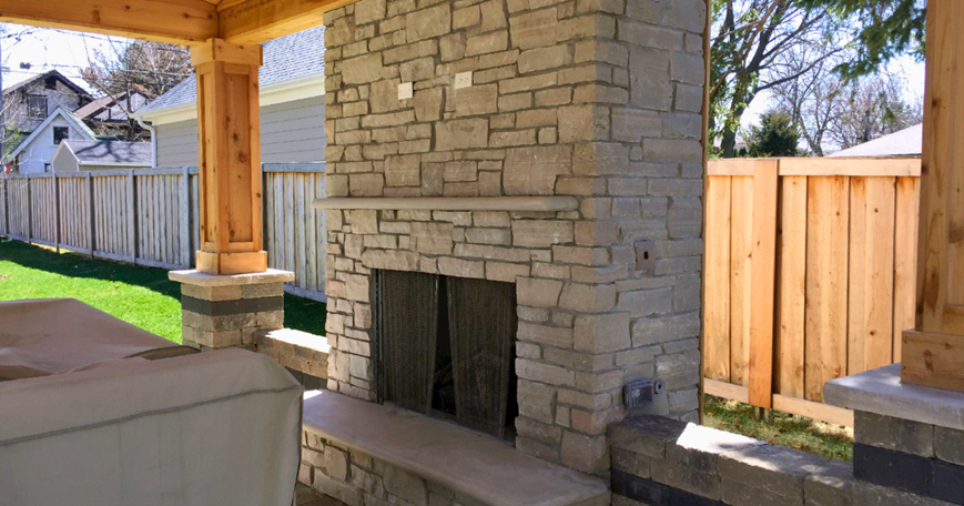 Luxurious custom outdoor stone fireplace with chimney, built-in grill station, and patio seating on an interlocking paver patio in the evening.