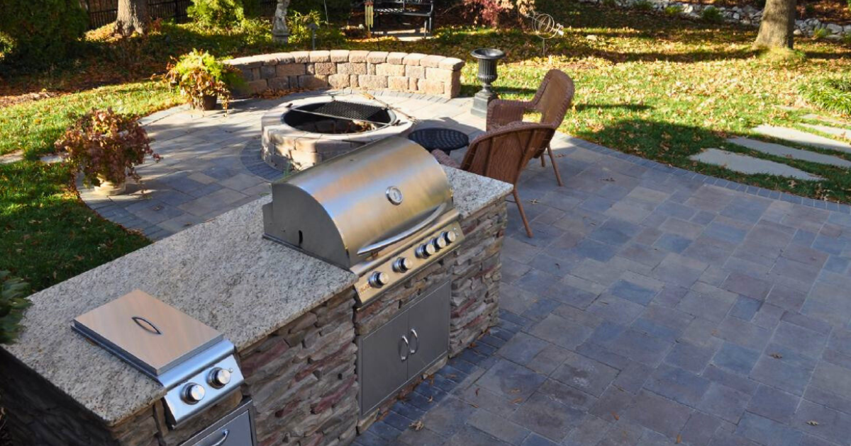 Integrated outdoor kitchen with granite countertop, built-in grill, stone base, and adjacent custom stone fire pit on a paver patio.