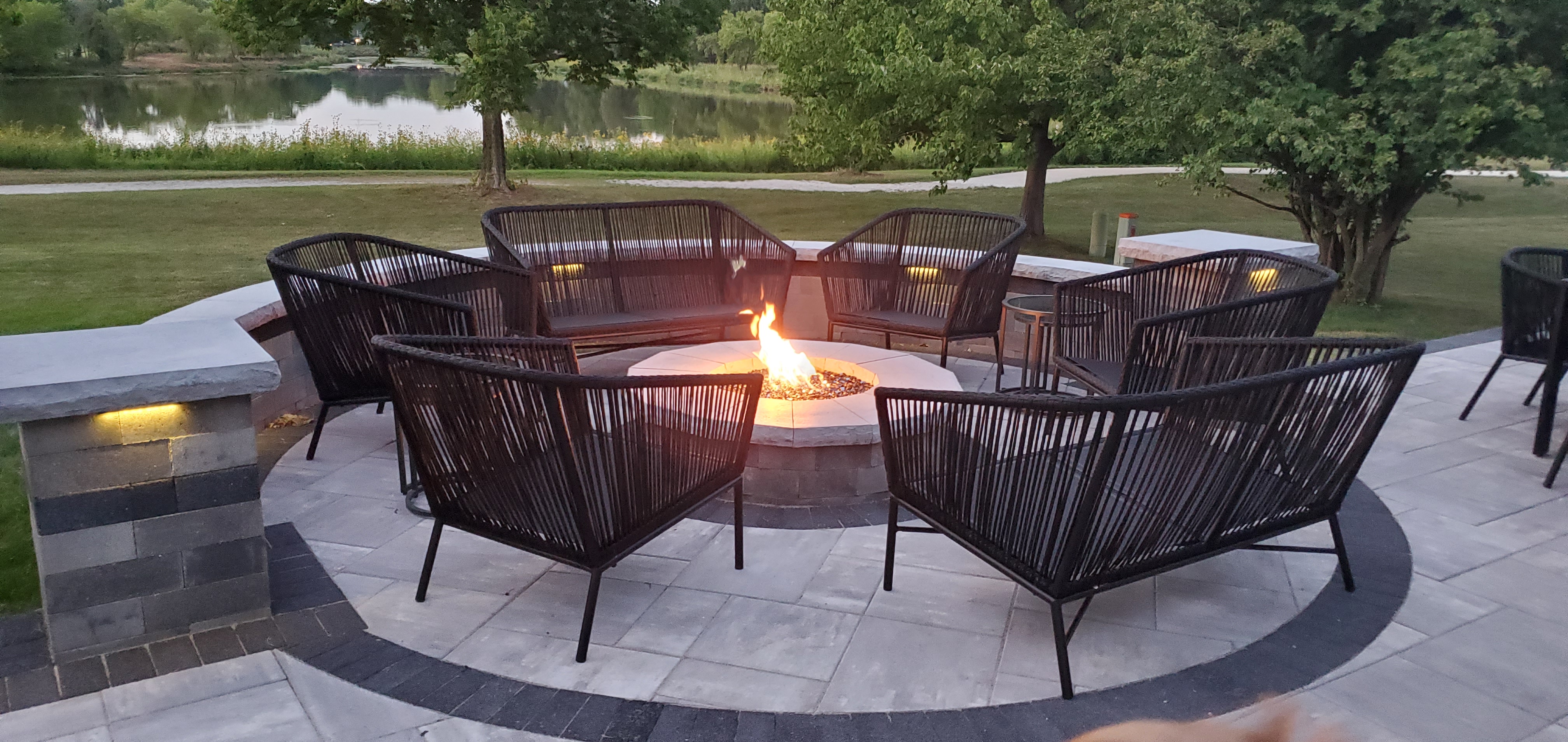A large, round fire pit, a custom fire feature, on a paver patio with modern chairs.