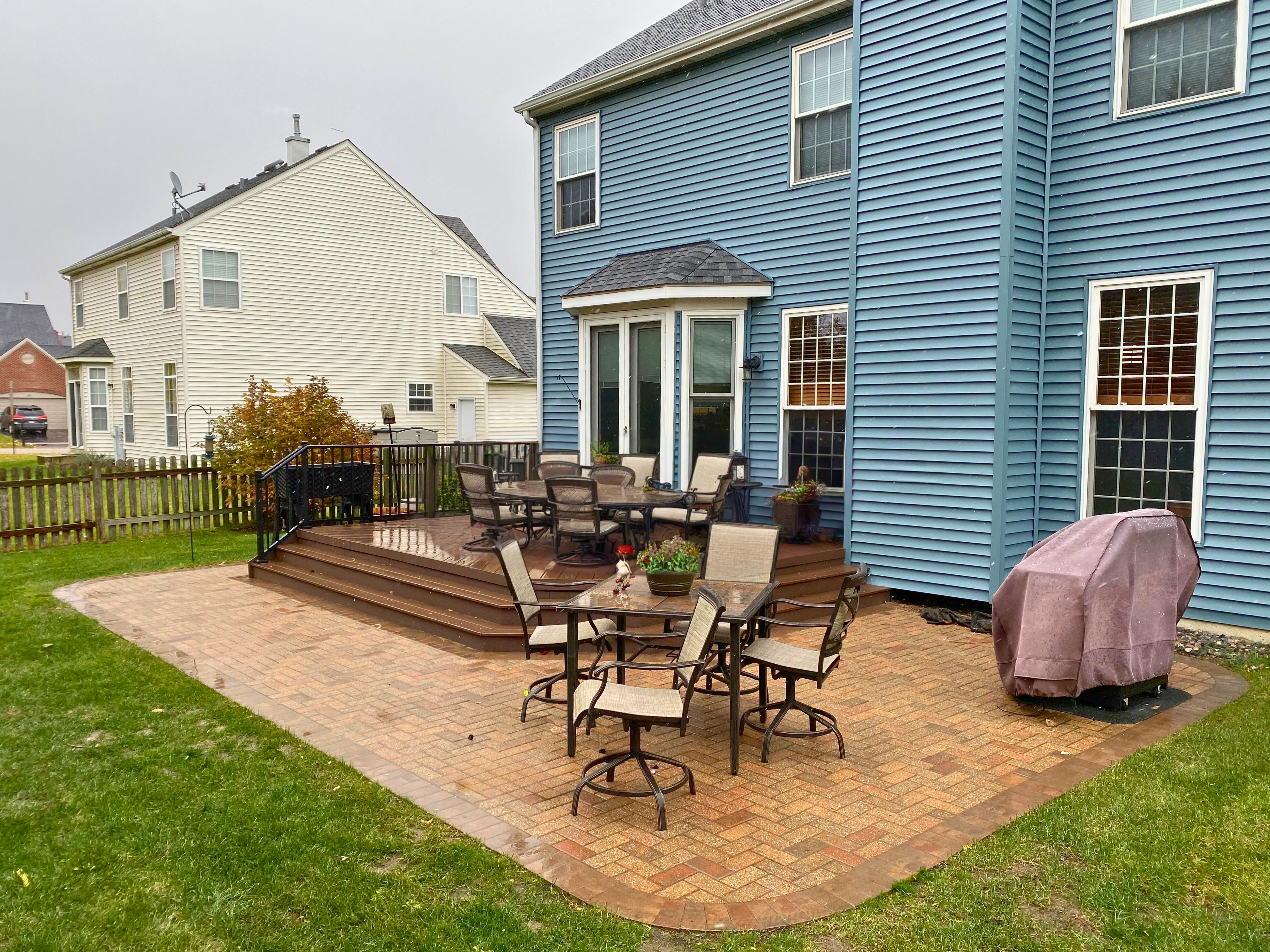 A multi-level outdoor living space featuring a reddish-brown paver patio with a dark border, connected to a composite deck with black railings, extending from a blue siding house.