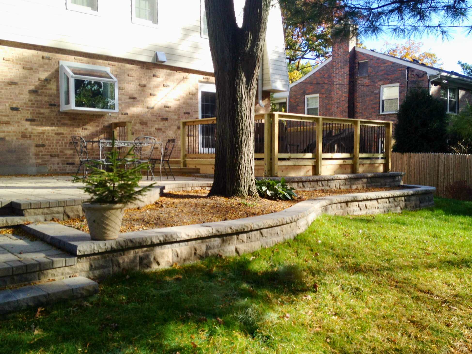 A multi-level outdoor living space in Lake Forest, featuring a natural wood deck with black metal balusters connected to a brick home, and an adjacent stone paver patio with a curved retaining wall and steps, surrounding a large tree.