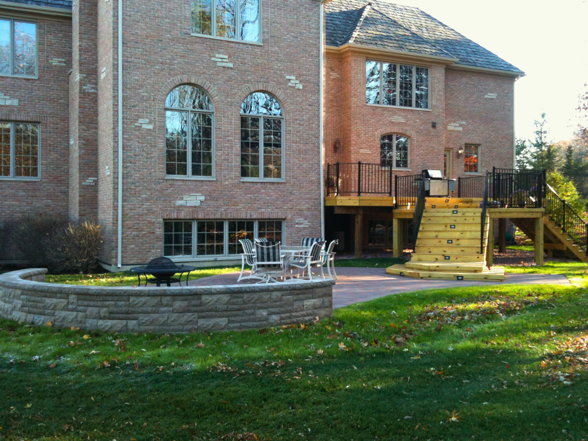 Deck and patio combination in Mundelein, IL, featuring a raised wood deck with black railings, a paver patio with seating, and a stone fire pit, next to a brick house.