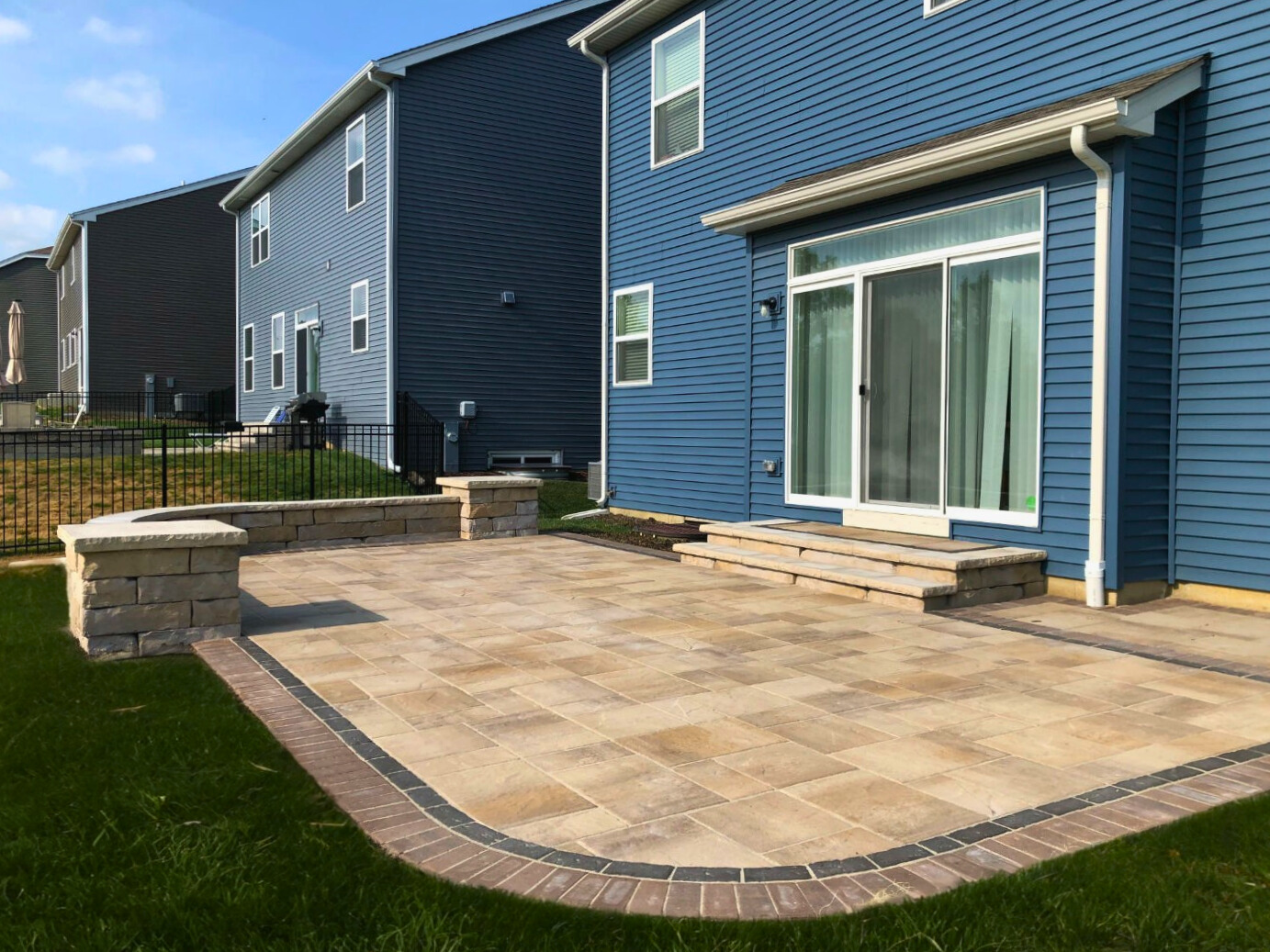 Modern raised paver patio in Gurnee with stone retaining walls, providing ample outdoor living space next to a blue house.