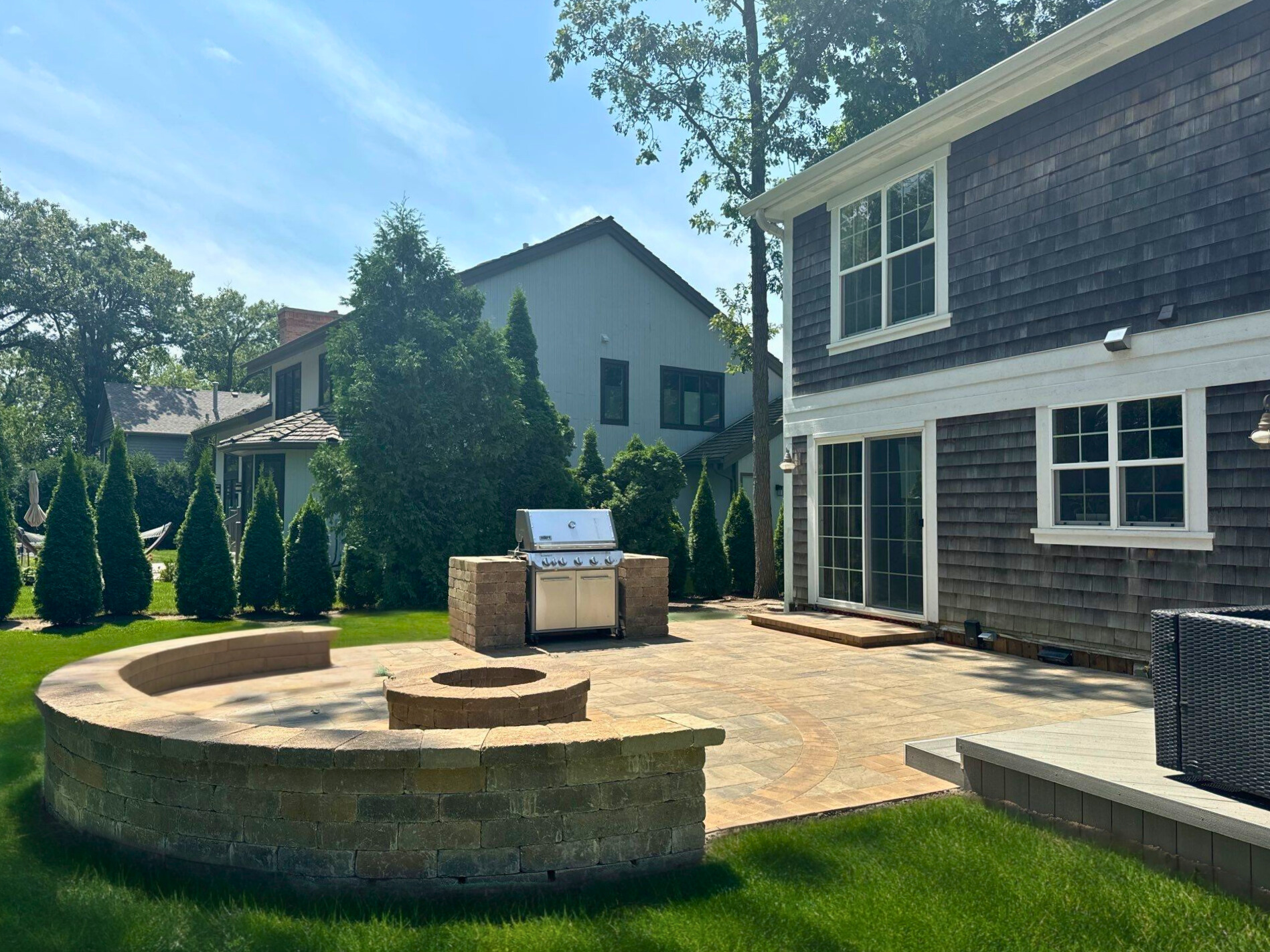 A spacious and intricately designed stone paver patio featuring a curved built-in seating wall, a grill station, and a fire pit, next to a modern shingle-sided home with large windows and a small composite deck section, all set in a sunny Lake Forest backyard.