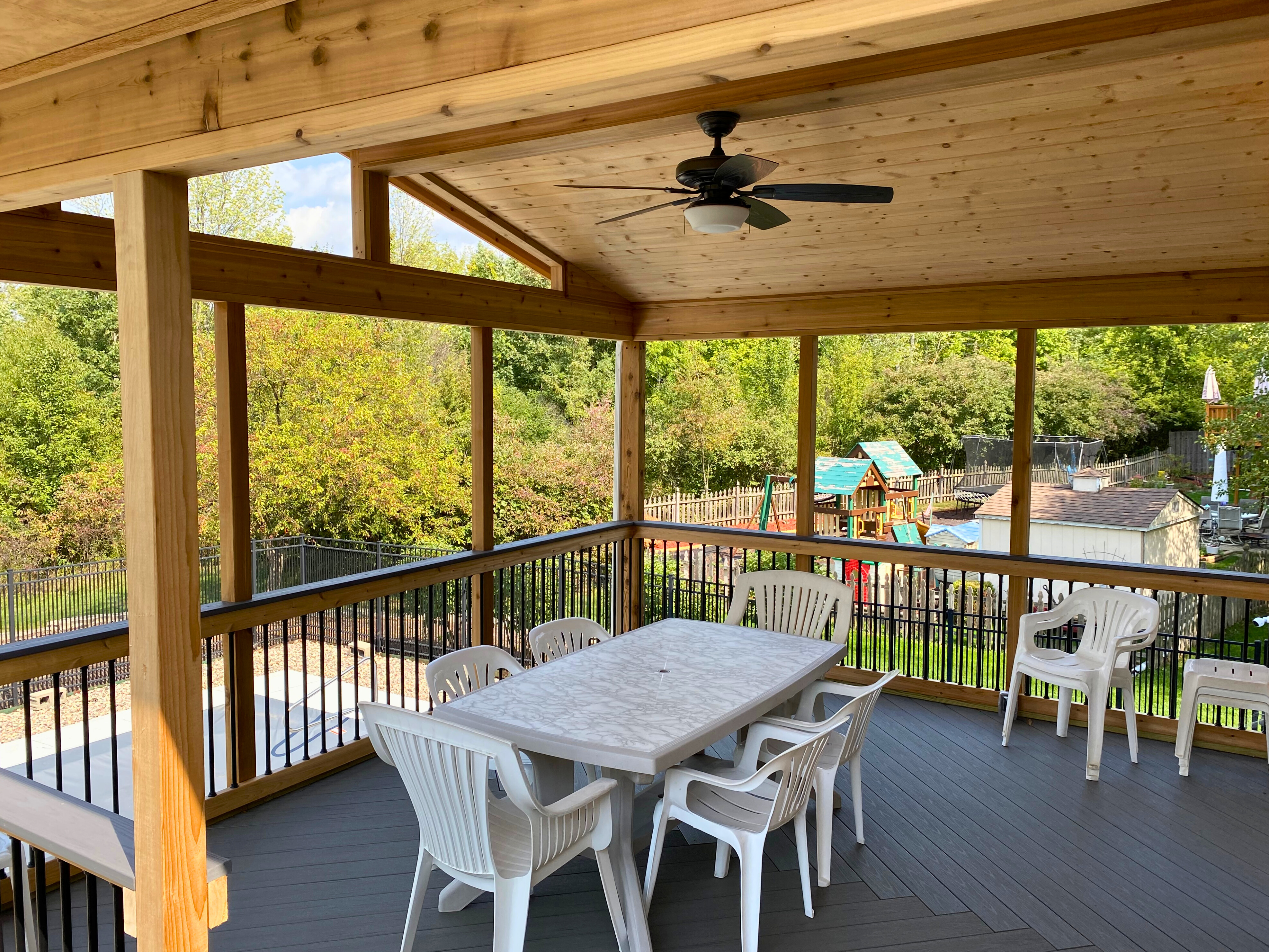 The interior of a spacious open porch with a light wood ceiling and frame, a ceiling fan, and grey composite decking, featuring a white outdoor dining set and overlooking a backyard with a playground.