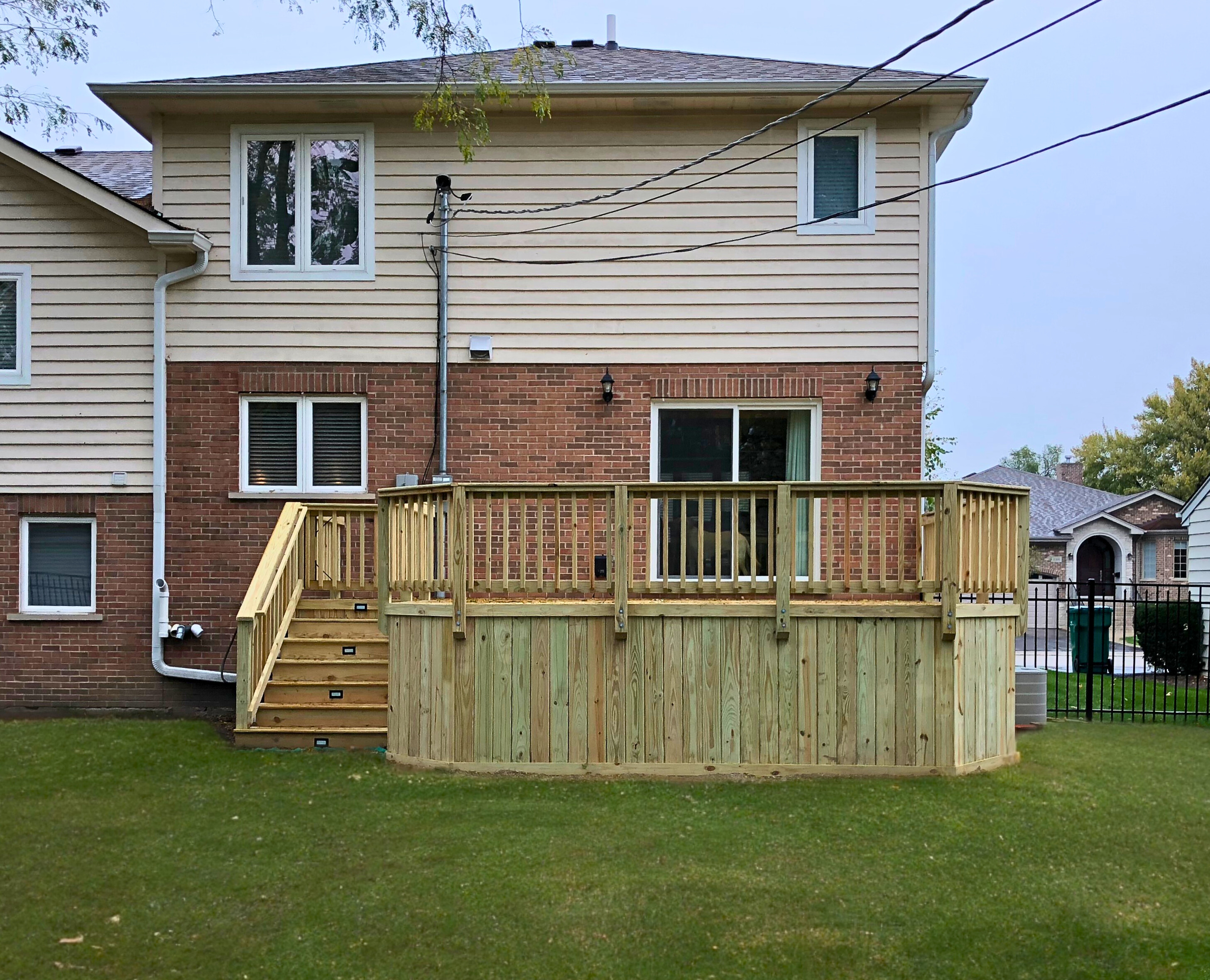 A newly built custom wood deck with stairs, a railing, and a privacy skirt, attached to a two-story home with brick and siding, overlooking a green lawn in Deerfield, IL.