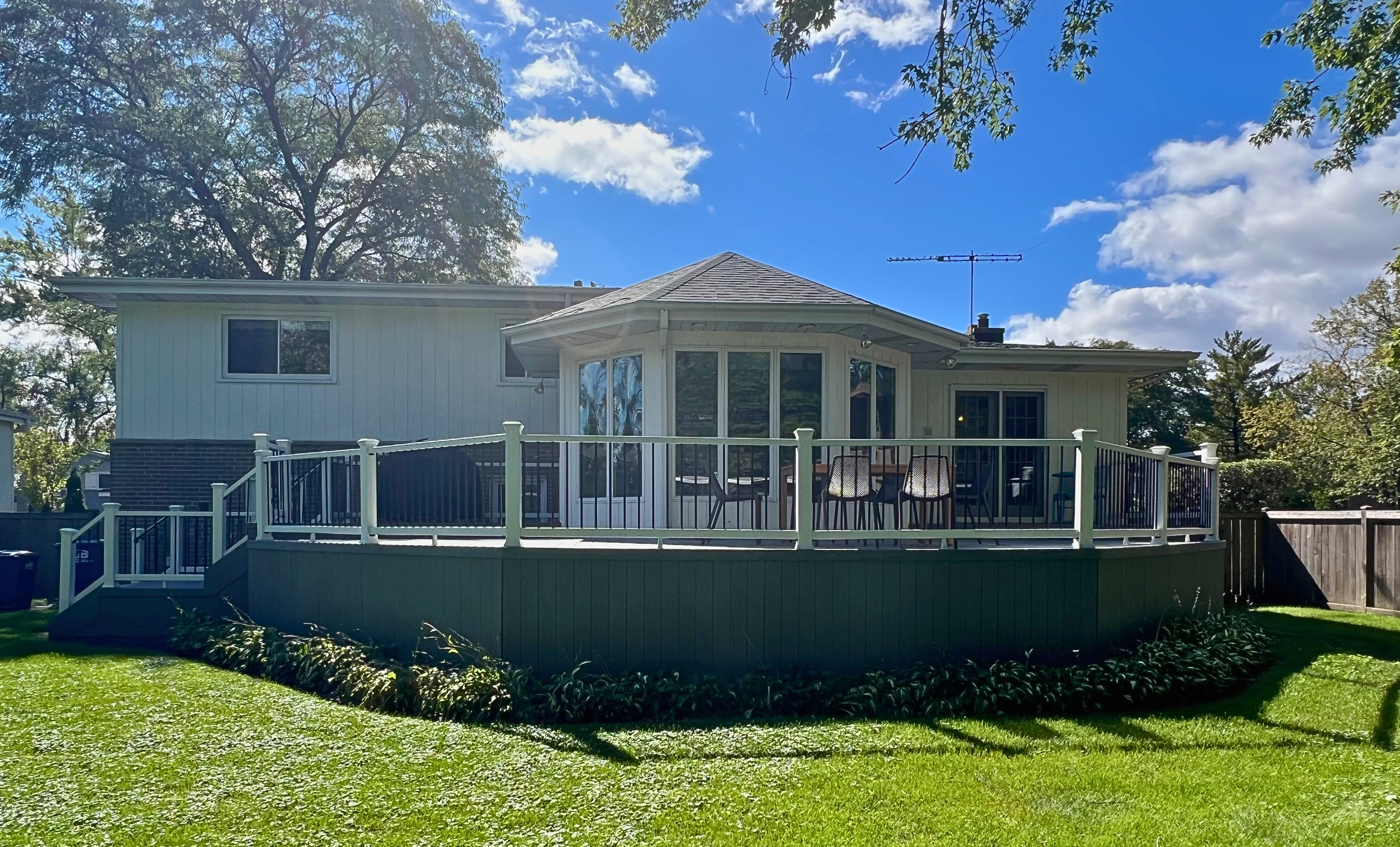 : A curved composite deck with white railings and black balusters, attached to a light-colored home with a prominent bay window, overlooking a lush green lawn in Deerfield, IL, under a bright blue sky.