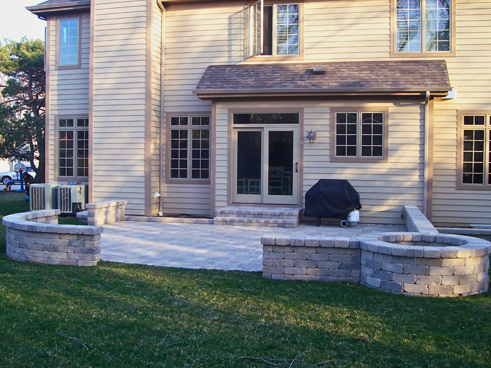 A spacious stone paver patio with curved retaining walls, a built-in fire pit, and a grill area, attached to a light-colored home with French doors, in a sunny Deerfield, IL backyard.