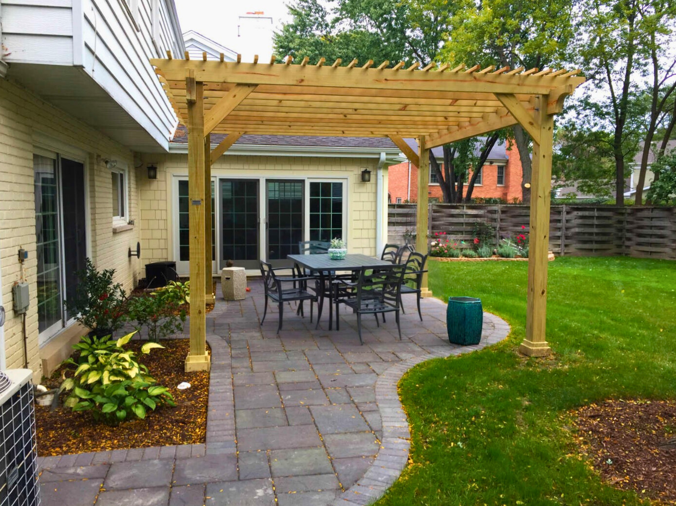 A spacious stone paver patio with a dining set, covered by a newly built wooden pergola, next to a light-colored brick home in Deerfield, IL, with a green lawn.