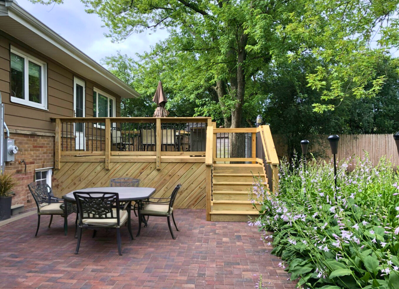 A natural wood deck with black metal balusters and matching stairs leading down to a brick paver patio with outdoor dining furniture, surrounded by lush plants and a wooden fence, in Deerfield, IL.