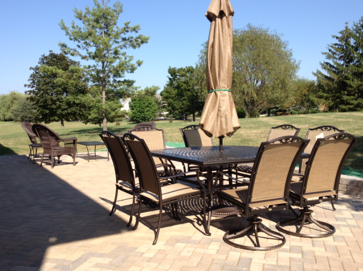A large paver patio furnished with an outdoor dining set and umbrella, surrounded by green landscaping and trees under a clear blue sky in Hawthorn Woods, Illinois.